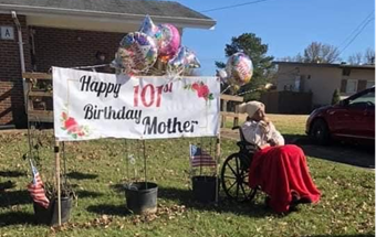 Ms. Bradford sits next to a large banner that reads Happy 101st Birthday Mother with ballons and flower pots